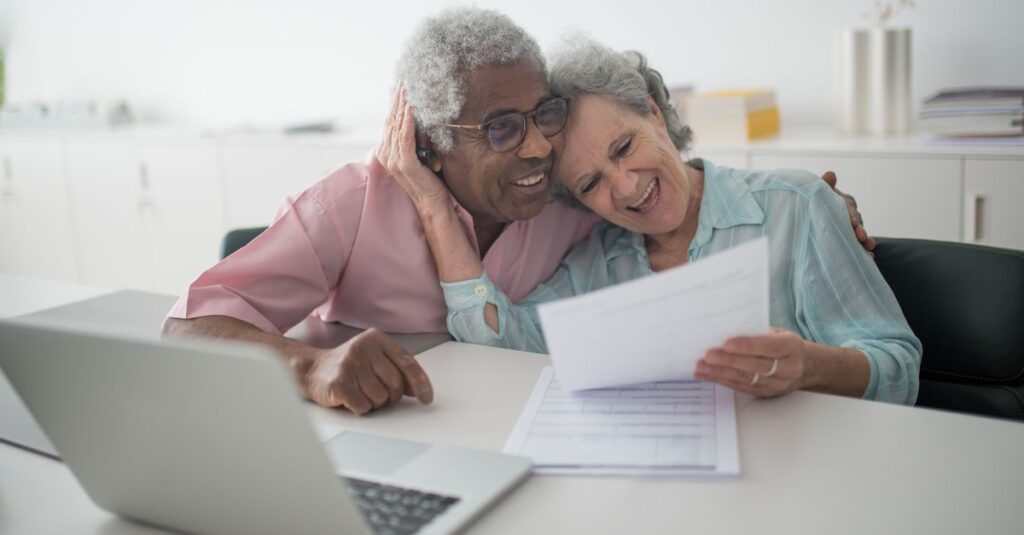 A joyful senior couple smiling while reviewing documents together at a desk with a laptop.
