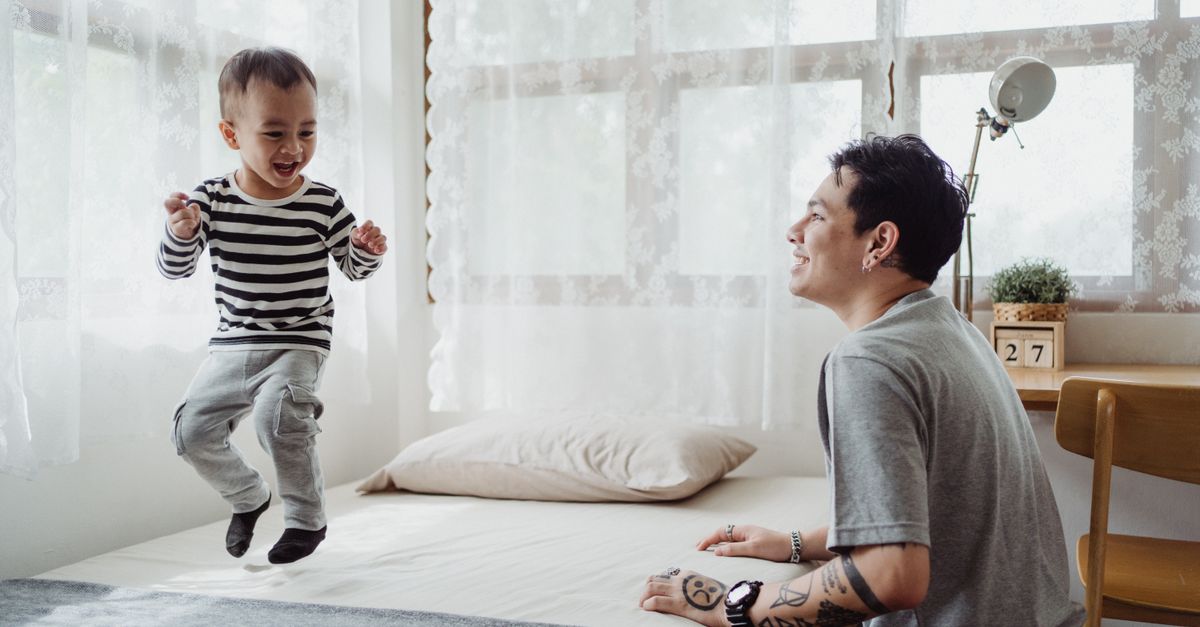 A joyful father watches his son jump on a bed in a bright, cozy bedroom.