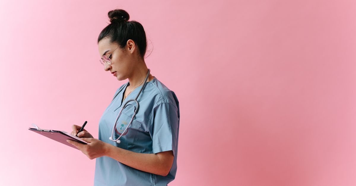 Female nurse in scrubs writing on a clipboard with a stethoscope on a pink background.