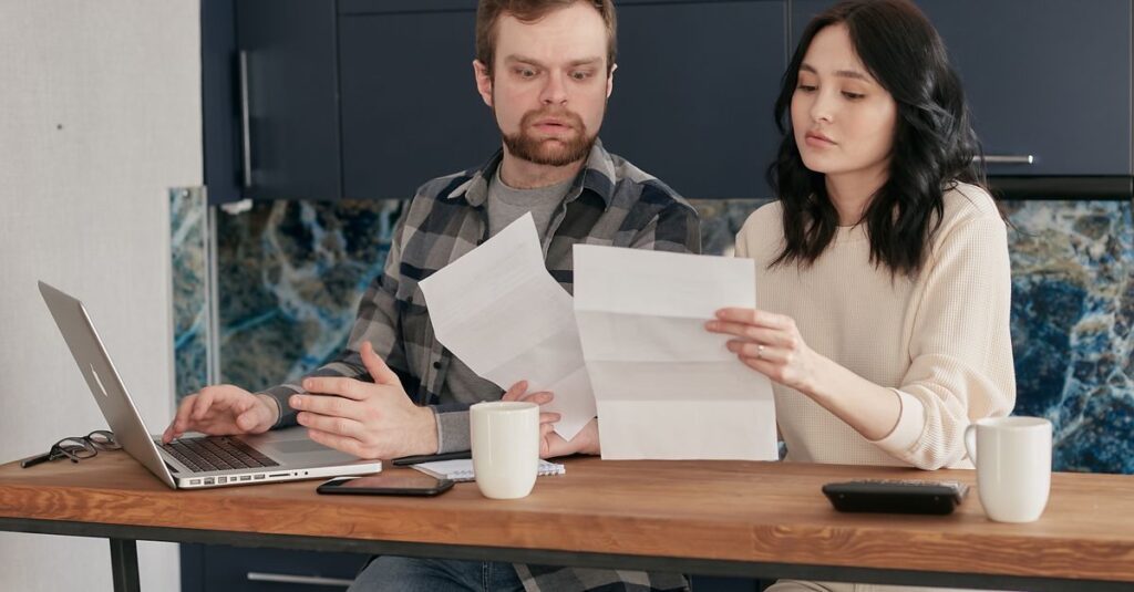 A couple sitting at a wooden table, reviewing documents with a laptop nearby, expressing concern.