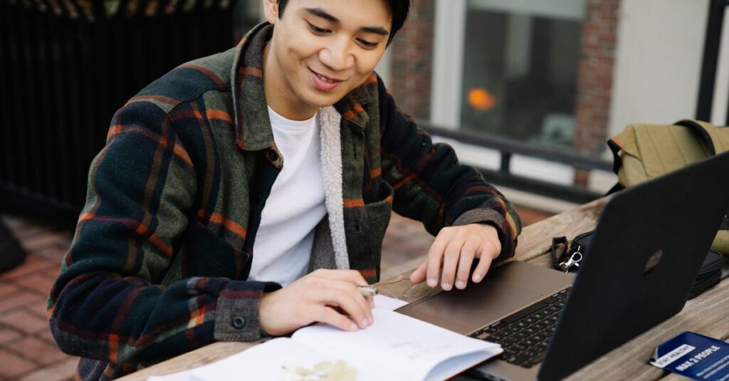 Adult male studying outside using a laptop and a notebook on a wooden table. Education concept.